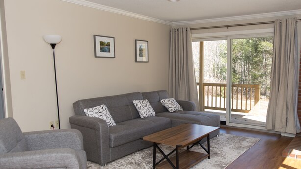 Ein Wohnzimmer mit grauem Sofa, einem Holztisch und einem gemusterten Teppich. An der Wand hängen zwei gerahmte Bilder und es steht eine Stehlampe.