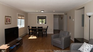Ein Wohnzimmer mit Flachbildfernseher, grauem Sofa, Esstisch mit Stühlen und einem Fenster mit Jalousien.
