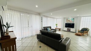 Un salón amplio con un sofá negro grande, un televisor y una ventana con cortinas traslúcidas.