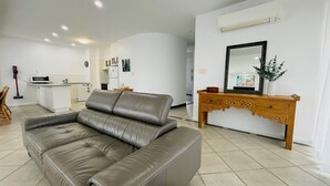 Un salón moderno con un sofá de cuero gris, una mesa consola de madera con un espejo y una cocina al fondo.