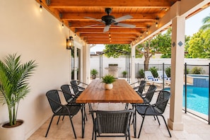 An outdoor patio with a wooden table and chairs, a ceiling fan, string lights, and a pool in the background.