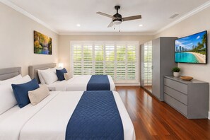 A bedroom with two beds, a ceiling fan, a TV, and a view of palm trees through the window.