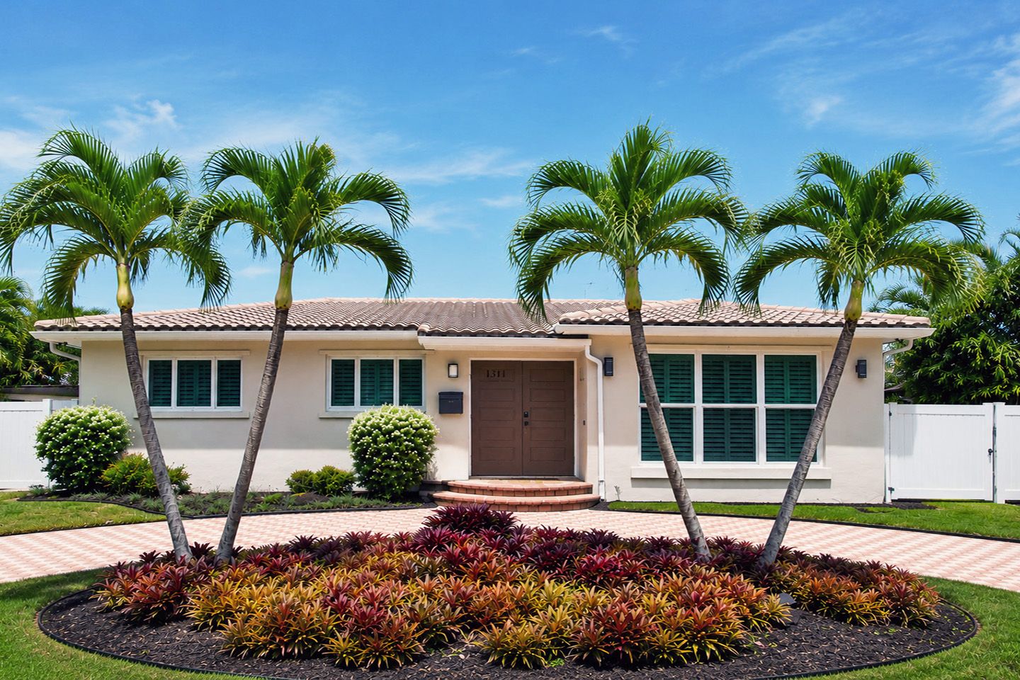 A two-story house with a tiled roof, a front door, and a small front yard with palm trees and a flower bed.