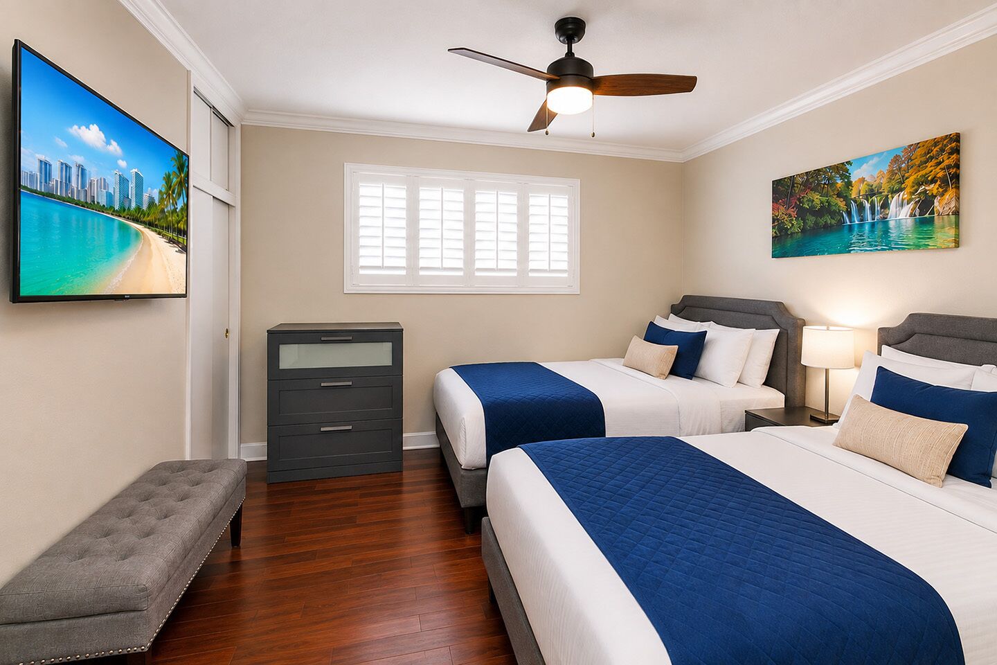 A hotel room with two beds, a desk, a bench, a ceiling fan, and a window with blinds.