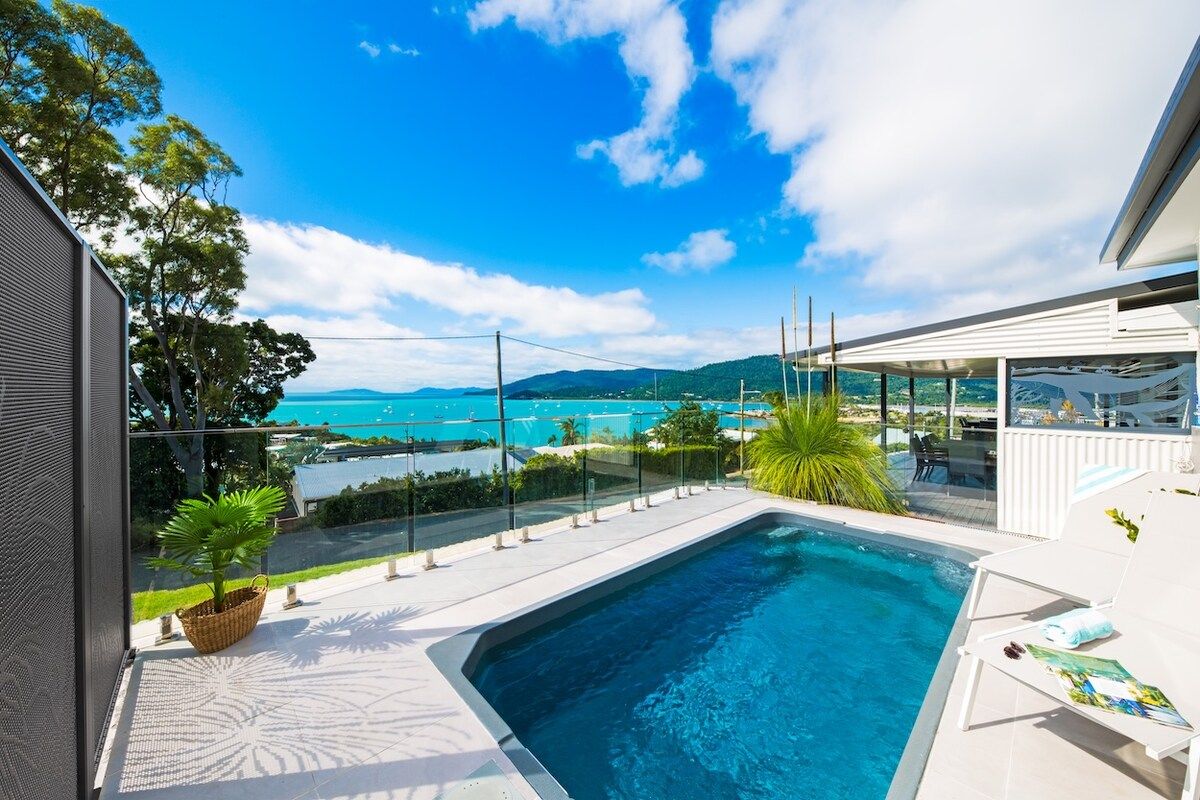 A swimming pool with a view of the sea and mountains, surrounded by a covered patio area.