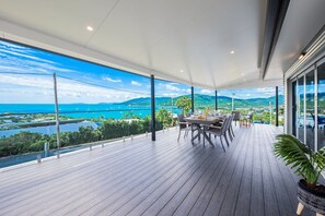 Un balcón amplio con piso de madera, una mesa de comedor y sillas, con vistas panorámicas a una masa de agua y colinas.