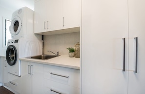 A modern kitchen with a built-in washer and dryer, stainless steel sink, and white cabinetry. - Airlie Blue On Blue (Airlie Beach)