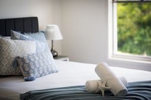 A neatly made bed with patterned pillows, a rolled towel with a starfish, and a lamp on a bedside table. - Airlie Blue On Blue (Airlie Beach)