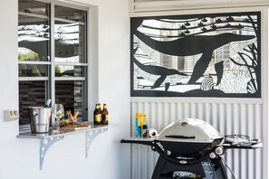 A room with a window, a wall-mounted artwork, a motorcycle, and a small shelf with bottles and a glass. - Airlie Blue On Blue (Airlie Beach)