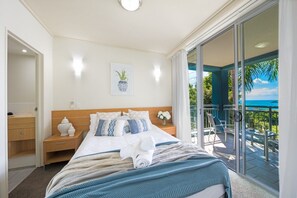 A hotel room with a bed, a nightstand, a window with a view of the outdoors, and a balcony with chairs. - Coastal Charm On Airlie (Airlie Beach)