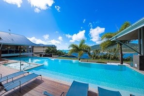 A swimming pool with lounge chairs and a deck, surrounded by palm trees and a view of the sea. - Coastal Charm On Airlie (Airlie Beach)