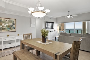 A spacious living area with a wooden dining table, a sofa, and a large window offering a view of the ocean.