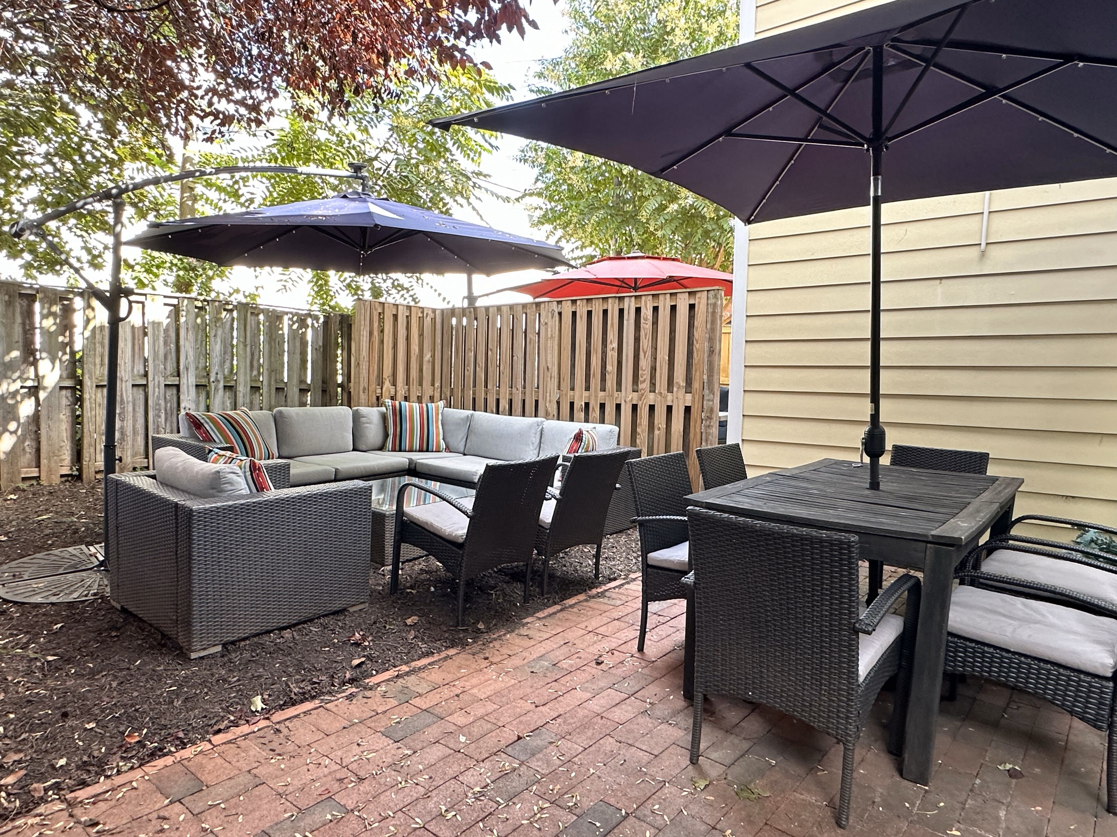 A patio with a sectional sofa, dining table, and chairs, surrounded by a wooden fence and covered by two umbrellas.