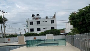 Un espacio al aire libre pavimentado con piscina, entrada protegida por un portón y un edificio de varios pisos al fondo.