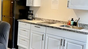 A compact kitchen with white cabinets, a granite countertop, a stainless steel refrigerator, a microwave, a coffee maker, and a sink with a faucet.