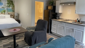 A compact hotel room with a bed, a small table, a chair, a sofa, a kitchenette, and a bathroom visible through an open door.