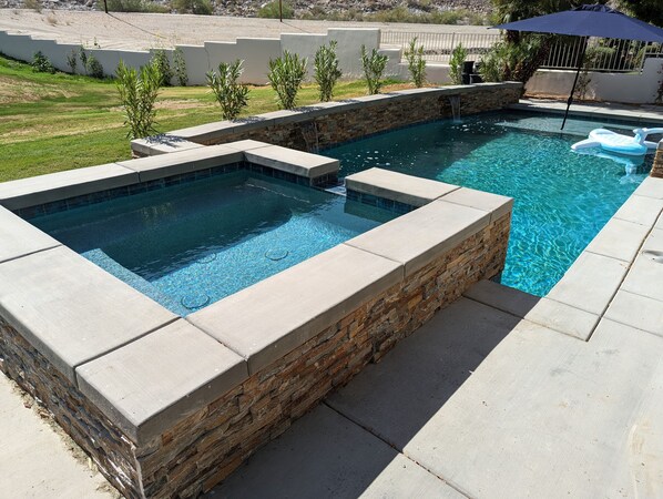 A rectangular swimming pool with a stone and concrete border, surrounded by a patio area with a lawn and a fence in the background.