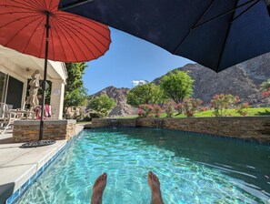 Ein Pool mit klarem Wasser, ein roter Sonnenschirm und ein Berg im Hintergrund.