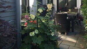 Een terras met een tafel, stoelen, een rode parasol en verschillende planten.