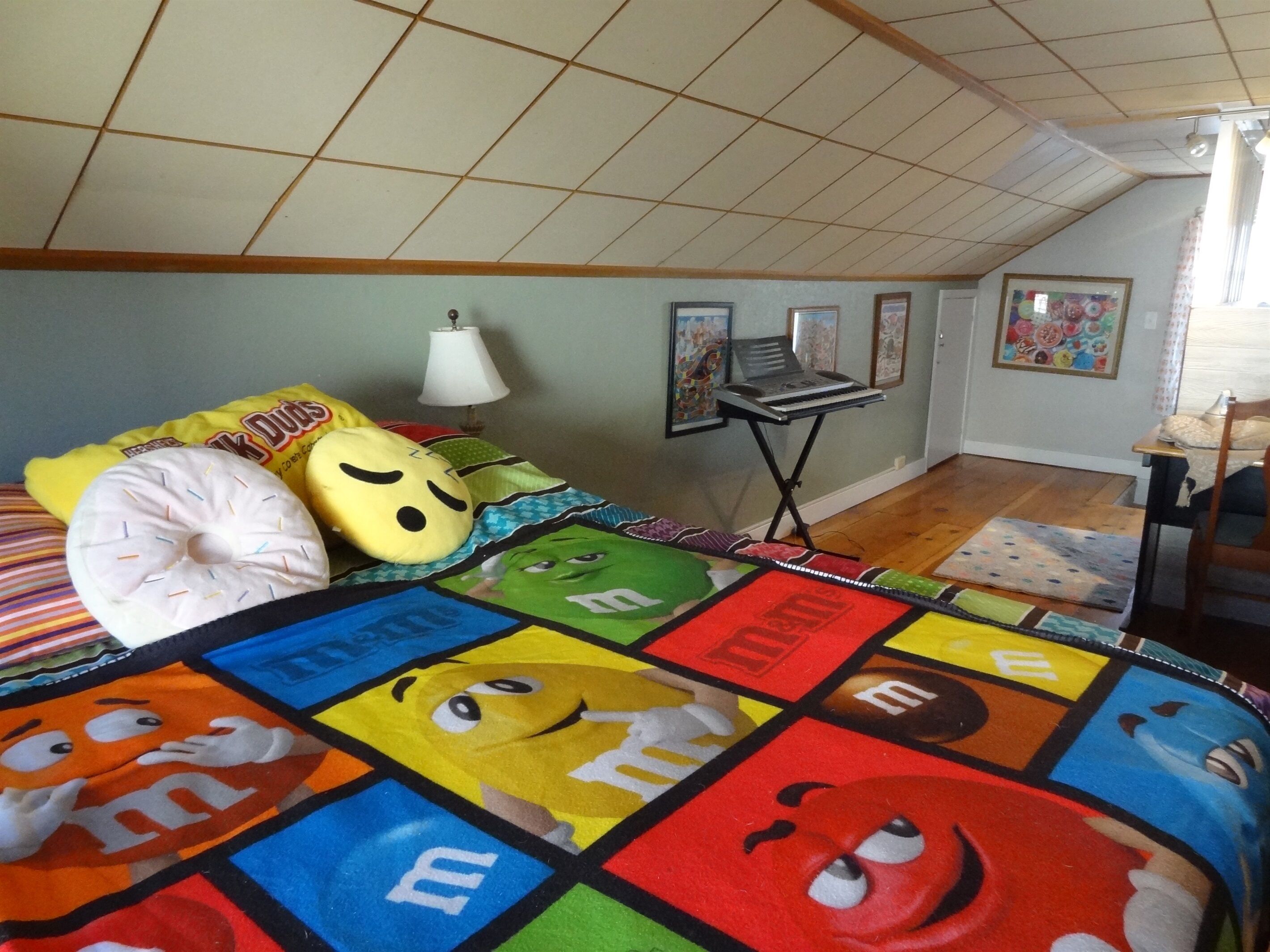 Un dormitorio con techo alto, una cama con ropa de cama colorida, un escritorio con lámpara y cuadros en la pared.