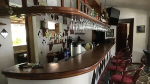 Eine Bar mit einer gebogenen Holztheke, zahlreichen Flaschen, Gläsern und einer Kaffeemaschine. Rote gepolsterte Stühle und ein strukturierter Boden sind ebenfalls vorhanden.