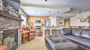 A modern living room with a stone fireplace, leather sofa, and a kitchen in the background. - Ski-In/Ski-Out Granby Base Camp Escape w/ Balcony (Granby)