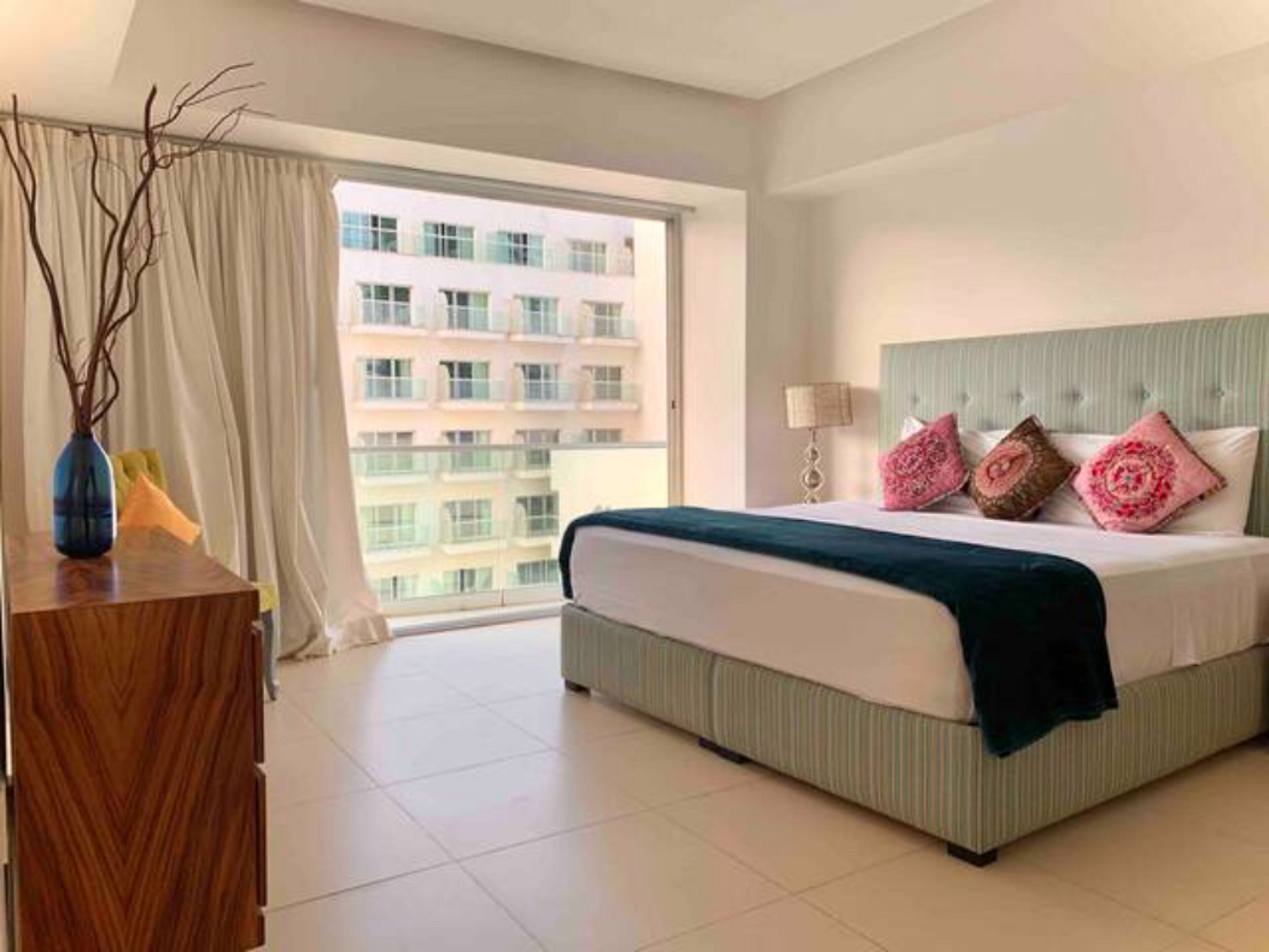 A hotel room with a large bed, a nightstand, a lamp, and a view of a building through the window.