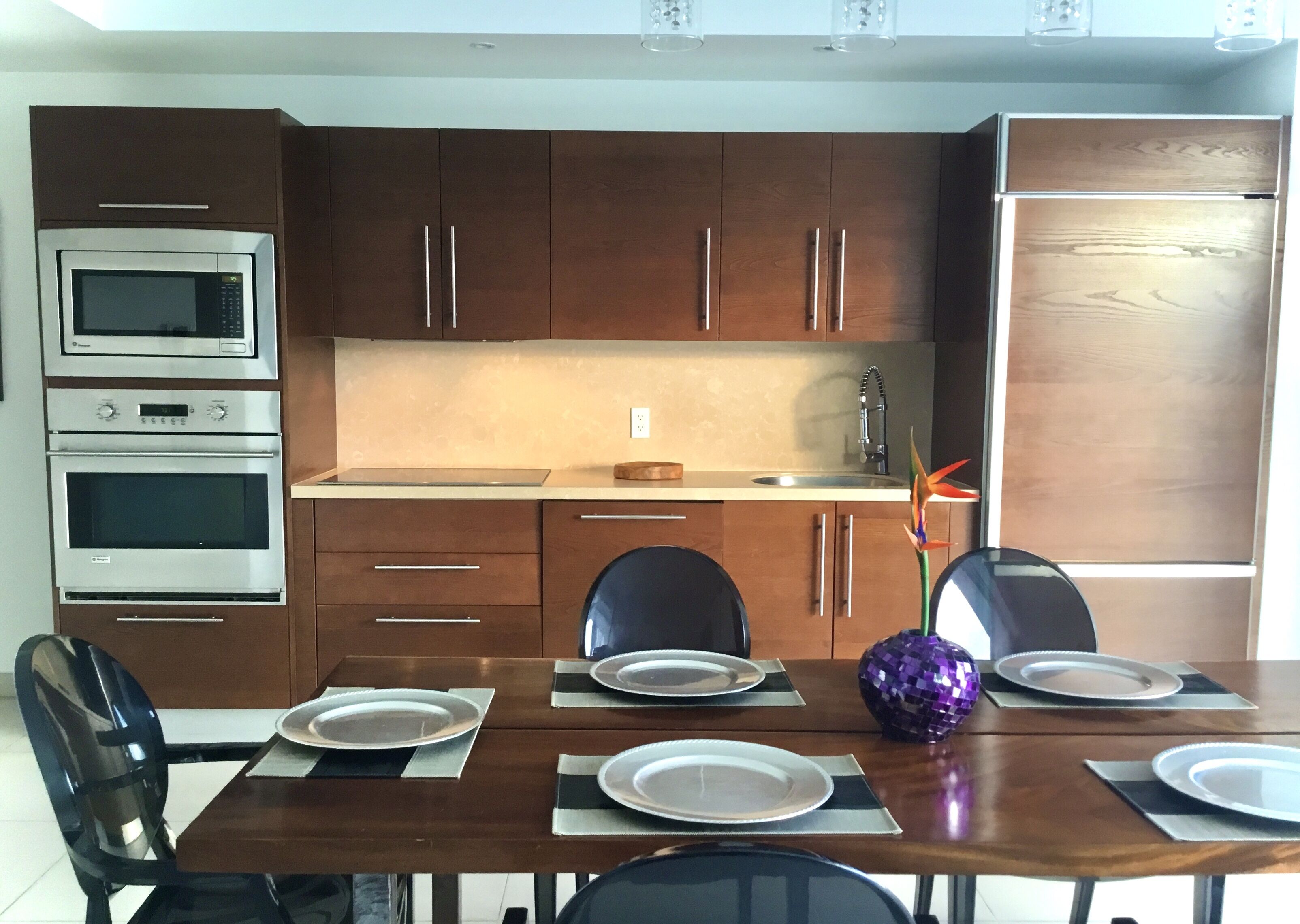 A modern kitchen with wooden cabinets, stainless steel appliances, a dining table set for four, and a decorative plant.