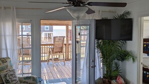 Ein Zimmer mit Deckenventilator, einem an der Wand befestigten Fernseher, einer Holzterrasse mit Gartenmöbeln und Blick aufs Meer.