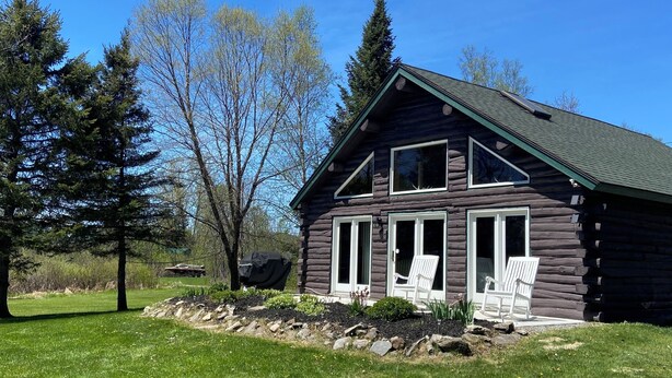 Une cabane en rondins dotée d’une grande fenêtre, d’un porche abrité et d’une cour gazonnée.
