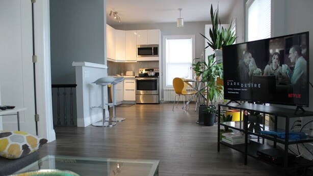 A modern living room with a flat-screen TV, a glass coffee table with fruit, and a kitchen in the background.