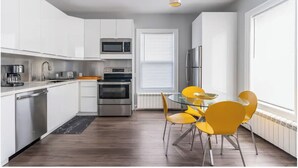 A modern kitchen with white cabinets, stainless steel appliances, a glass dining table with yellow chairs, and a window with blinds.