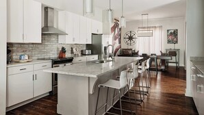 Uma cozinha moderna com armários brancos, uma ilha central com banquetas, uma janela grande com cortinas e uma peça de arte de parede decorativa com uma bússola.