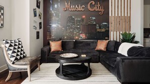 Uma sala de estar moderna com um sofá preto, uma mesa de centro redonda e uma obra de arte na parede retratando um horizonte urbano, identificada como "Music City".