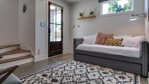 Een moderne woonkamer met een grijze bank, een kamerbreed tapijt met patroon, een houten trap en een raam met uitzicht op groen.