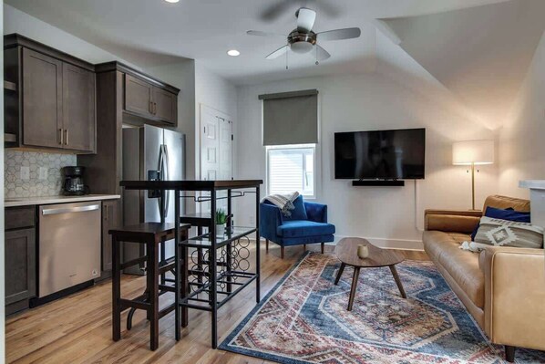 Une cuisine moderne avec des meubles sombres, un réfrigérateur en acier inoxydable et un coin repas avec une table et des chaises en bois. Le salon est doté d’un canapé bleu et d’un divan en cuir marron, et un téléviseur est fixé au mur.