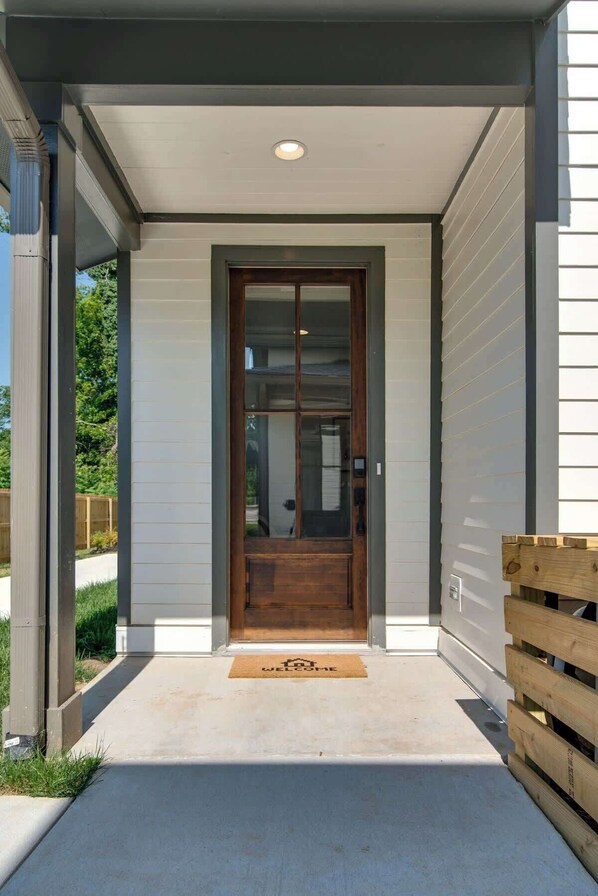 A modern house entrance with a wooden door, a welcome mat, and a concrete pathway leading to the door.