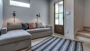 Ein modernes Wohnzimmer mit einem grauen Ecksofa, zwei Stehlampen, einem gemusterten Teppich und einem großen Fenster mit Jalousien.