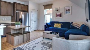 A modern living room with a blue sofa, a coffee table, and a kitchen area with wooden cabinets and a built-in refrigerator.