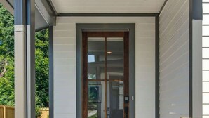 A modern house entrance with a glass door, a wooden door, and a concrete pathway leading to the door.