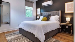 A modern bedroom with a large bed, a bedside table, a wall-mounted air conditioner, and a window with a view of greenery.