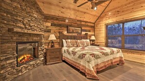 Une chambre confortable dans un chalet en rondins, avec une cheminée en pierre, un lit recouvert d’une couverture à motifs, des murs en bois et une grande fenêtre.