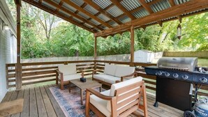 Un patio exterior con mobiliario de madera, una parrilla y una zona cubierta con vistas al jardín trasero.