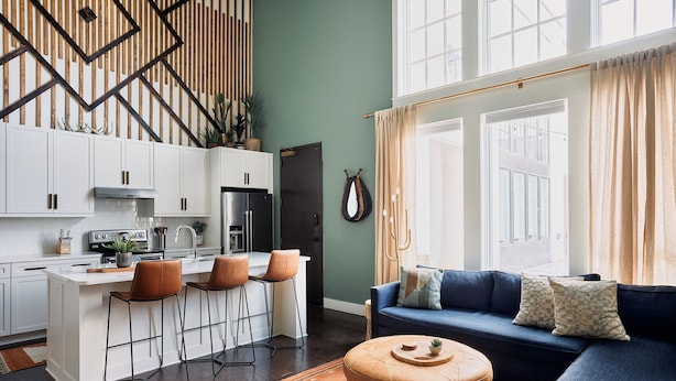 A modern kitchen with white cabinets, a central island with bar stools, and a blue sofa with patterned cushions in a living area with large windows and curtains. - Stay In The Heart Of 12 South, Designer Penthouse  |  1 of 7 Themed Lofts at Cowrite, Find Coffee and Tacos Downstairs, Complimentary Spin Class, Walk to Shopping in 12 South, 10 Minutes to Broadway, Parking, 24 Hr Hospitality (Nashville)
