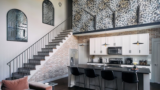 A modern kitchen with a central island, a staircase with a metal railing, and a brick wall. - Johnny Cash Inspired 2-Suite Loft In 12 South      |         1 of 7 Themed Lofts at Cowrite, Find Coffee and Tacos Downstairs, Complimentary Spin Class, Walk to Shopping in 12 South, 10 Minutes to Broadway, Parking, 24 Hr Hospitality (Nashville)