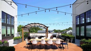 Una terraza en la azotea con una mesa y sillas de madera, rodeada de macetas y una pérgola.