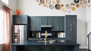 Una cocina moderna con armarios grises oscuros, una isla central con taburetes, un refrigerador de acero inoxidable y una variedad de platos tejidos en la pared.