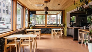 Un restaurant avec des tables et des chaises en bois, de grandes fenêtres et un espace cuisine visible grâce à l’aménagement ouvert.