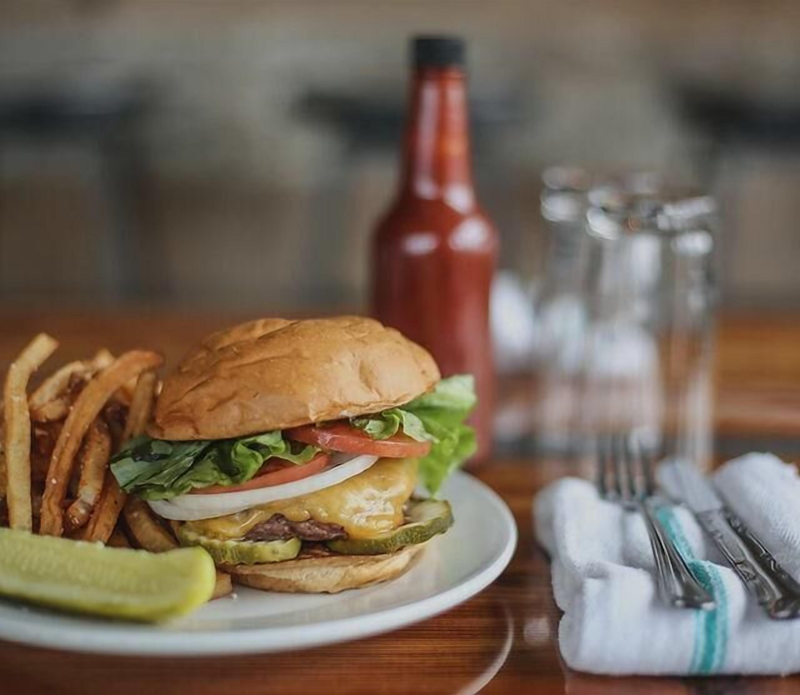 Hamburguesa con papas fritas y pepinillo en un plato, una botella de kétchup y vasos sobre una mesa.