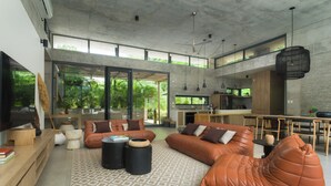 A spacious living area with leather sofas, a coffee table, and a large TV. There is a kitchen area with a dining table and chairs visible through an open space. - Casa Taman, Beachside villa by Elite Beach Villas (Cobano)
